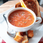 A bowl of vibrant roasted tomato basil soup, garnished with fresh basil leaves and mini croutons.
