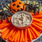 A colorful Halloween pumpkin veggie tray arranged on a platter, featuring carrots, black olives, bell peppers, and cucumbers.